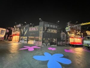 Nighttime scene with neon signs in Korean, colorful flower-shaped street art, and urban ambiance.