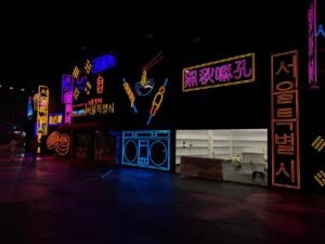 Nighttime street scene with neon signs in purple, blue, and red, advertising food and entertainment.