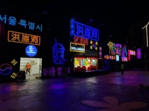 Nighttime street scene with neon signs in purple, blue, and red, advertising food and entertainment.