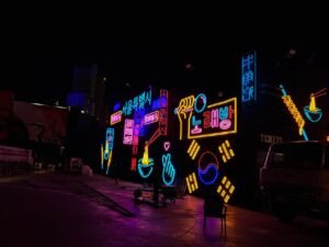 Nighttime street scene with neon signs in purple, blue, and red, advertising food and entertainment.