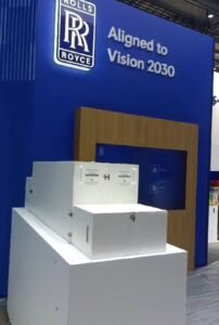 Blue and white Rolls-Royce exhibit display with 'Aligned to Vision 2030' text, wooden panel, and stacked boxes