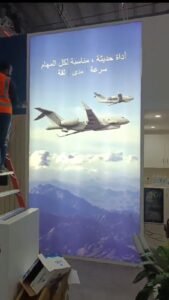 Worker on ladder beside illuminated poster with airplanes and Arabic text one exhibitions and events